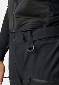 Nahaufnahme von schwarzen Hosen mit doppelten Druckknöpfen, Reißverschlusstasche, Stoffgürtel, D-Ring-Befestigung und Teil eines schwarzen Langarm-Tops.