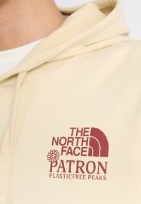 Sweat à capuche beige avec logo The North Face bordeaux, une fleur souriante et le texte "PATRON PLASTICFREE PEAKS" sur la poitrine.