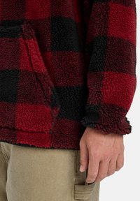 Red and black plaid fleece pullover with a textured surface, featuring a kangaroo pocket and an adjustable hem at the cuffs.