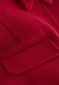 Nahaufnahme eines roten Wollstoffs mit einer flachen Tasche und gestickten Kanten sowie einer Schlaufe darüber. Glatte Textur, leuchtende Farbe.