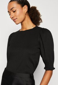 Black textured blouse with puff sleeves and gathered cuffs. Round neckline and a smooth, flowing appearance. Paired with a shiny black skirt.
