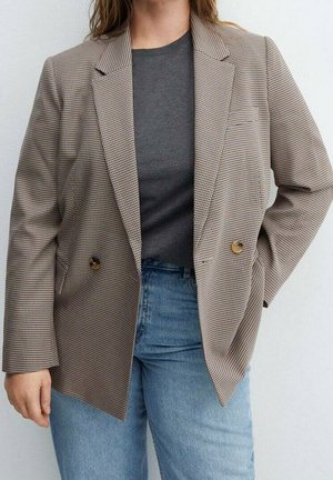 Vrouw draagt een bruin geruite blazer over een donkergrijze top met lichtblauwe jeans, staand met één hand in haar zak tegen een effen achtergrond.
