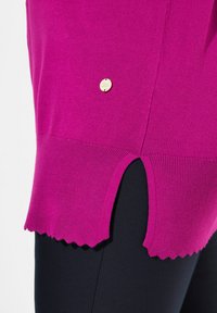 Fuchsia Pullover mit gewelltem Saum, der ein rundes goldenes Logo-Detail an der Seite und eine gerippte Textur am unteren Rand aufweist.