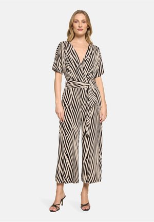 Femme portant une combinaison portefeuille à manches courtes, jambes larges et ceinture nouée à la taille, beige rayée de noir façon zèbre, accompagnée de sandales à talons noires.