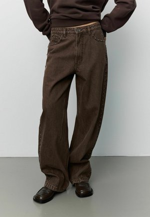Personne portant un jean large marron foncé avec des détails à clous sur les côtés et des chaussures marron foncé, debout devant un fond clair uni.