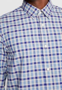 Camicia con bottoni a quadri, caratterizzata da quadrati in azzurro chiaro, blu scuro e viola. Tessuto di cotone con colletto standard e bottoni bianchi.