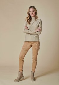 Beige, langärmliges Oberteil mit hohem Kragen und einem subtilen Schimmer, kombiniert mit hellbraunen Cargo-Hosen mit seitlichen Taschen und schmalem Schnitt. Graue Knöchelstiefel.