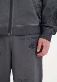 Completo sportivo grigio composto da giacca con zip e pantaloni. Il materiale è morbido, con design di tigri ricamati e scritte. Dettagli a coste sui polsini e in vita.