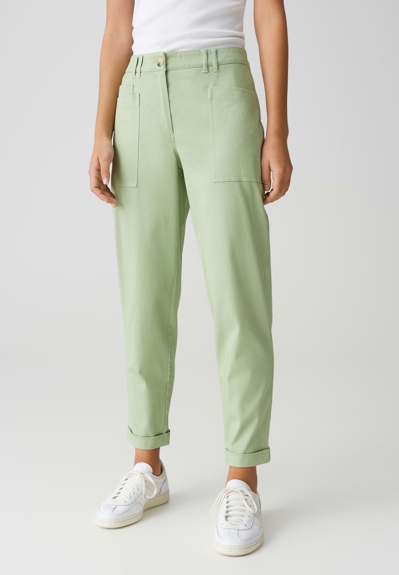 Personne portant un pantalon taille haute vert clair avec de grandes poches devant, des revers roulés, des baskets blanches et un haut blanc sans manches.