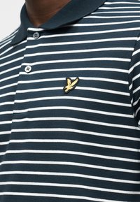 Πόλο πουκάμισο με οριζόντιες ρίγες σε ναυτικό μπλε και λευκό, με κίτρινο έμβλημα πουλιού στο αριστερό στήθος και δύο μαύρα κουμπιά στην πατιλέτα.