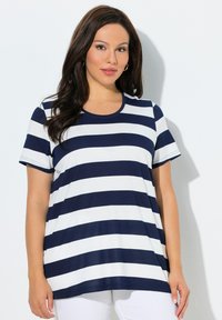 Frau mit langen dunklen Haaren, die goldene Creolen trägt und ein marineblau-weiß gestreiftes, kurzärmeliges Shirt sowie weiße Hose trägt.