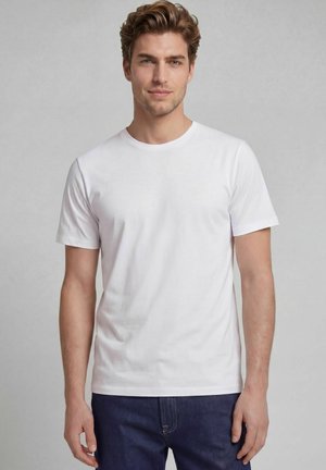 Jeune homme aux cheveux courts et ondulés portant un t-shirt blanc uni à manches courtes et un jean bleu foncé, debout devant un fond gris uni.