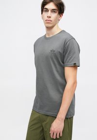 T-shirt cinza em algodão com gola redonda, mangas curtas e um pequeno logótipo no peito. Apresenta uma etiqueta de tecido em contraste na manga.
