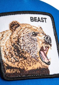 Gestickter Patch mit einem Bärenkopf in Brauntönen, umrandet von schwarz, auf einem weißen Hintergrund auf blauer Stoffbasis. Text lautet "BESTIE."