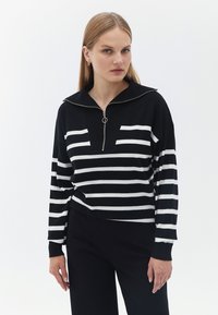 Schwarz-weiß gestreifter Strickpullover mit Reißverschluss am Ausschnitt, locker sitzend mit langen Ärmeln, vor einem schlichten weißen Hintergrund.