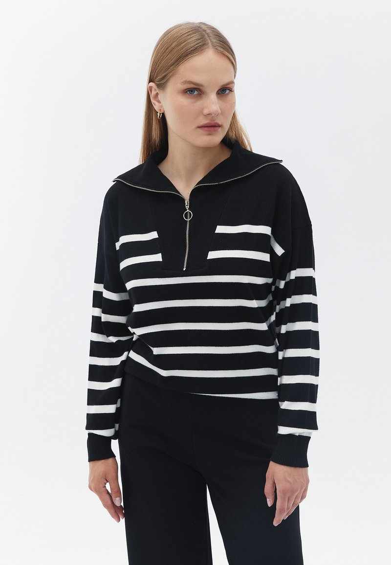 Schwarz-weiß gestreifter Strickpullover mit Reißverschluss am Ausschnitt, locker sitzend mit langen Ärmeln, vor einem schlichten weißen Hintergrund.