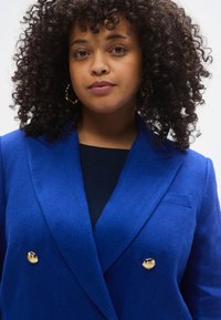 Plavi blazer s uzorkom zubaca psa s dvostrukim kopčanjem, zlatnim gumbima i džepom. Nošen preko tamnog vrha s teksturiranim završetkom.