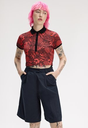Joven con cabello rosa brillante lleva un polo corto rojo con estampado de piñas y pantalones cortos negros holgados, con las manos en los bolsillos, sobre un fondo blanco.