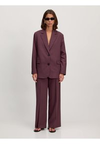 Lila maßgeschneiderter Anzug mit einem Blazer mit geknöpftem Revers und weit geschnittenen Hosen. Getragen mit schwarzen Sonnenbrillen und flachen Sandalen. Glatte Stofftextur.