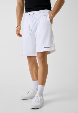 Uomo che indossa pantaloncini bianchi con coulisse e logo "GUESS JEANS", calzini bianchi, sneakers bianche e maglietta nera, mano in tasca.