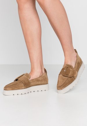 Füße in tanfarbenen Wildleder-Loafers mit großen Schnallendetails und dicken weißen Sohle auf einem glatten weißen Boden.