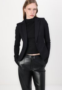 Black fitted blazer with a single button, paired with a black turtleneck and black leather pants featuring a zippered front. Smooth textures.