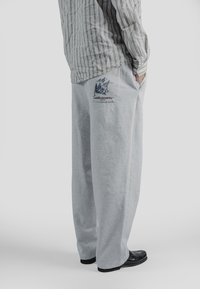 Osoba w luźnych szarych spodniach dresowych z nadrukiem gór i drzew na tylnej kieszeni, w paskowanej koszulce i czarnych butach, stojąca bokiem.