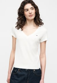 Γυναίκα με σγουρά μαλλιά που φοράει λευκό μπλουζάκι με V-λαιμόκοψη και σκούρο μπλε τζιν, κοιτάζοντας ελαφρώς προς τα πλάγια μπροστά σε απλή φόντο.