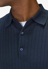Mörkblå stickad polo skjorta med vertikal ribbning, med en krage och två matchande knappar vid halsringningen.