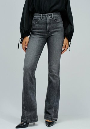 Personne portant un jean gris évasé à taille haute, un top noir transparent à manches longues et des escarpins noirs à talons pointus, se tenant devant un fond uni.