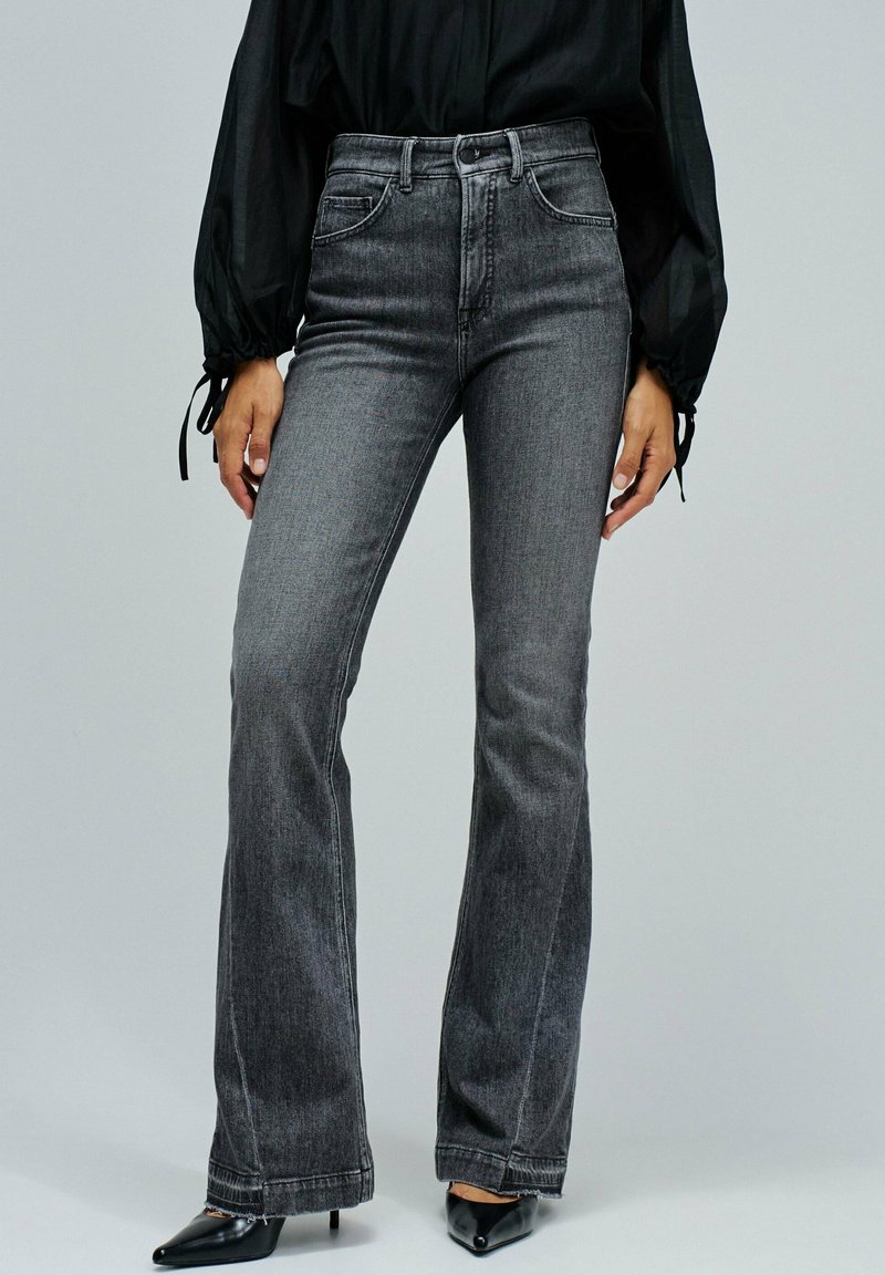 Osoba nosząca szare dżinsy z wysokim stanem w fasonie dzwonów, czarną przezroczystą bluzkę z długim rękawem oraz czarne szpilki z ostrym noskiem stoi na jednolitym tle.