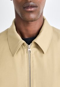 Chaqueta beige con cremallera y cuello, que presenta una cremallera plateada y una textura mate. El diseño enfatiza una apariencia limpia y moderna.