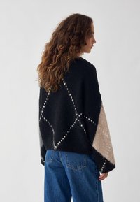 Kobieta z długimi kręconymi włosami, ubrana w czarny i beżowy sweter w wzory oraz niebieskie jeansy, stojąca bokiem na jednolitym tle.