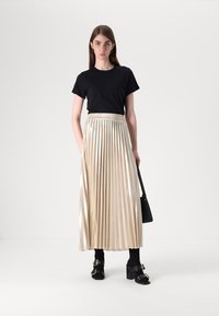 Un tricou negru asociat cu o fustă midi plisată, metalică aurie. Modelul poartă sandale cu toc negre și ține o geantă mică de mână neagră.