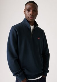 Donkerblauwe sweatshirt met een hoge kraag en een halve rits. Voorzien van elastische manchetbanden en een klein rood logo op de borst. Zacht, textuurstof.