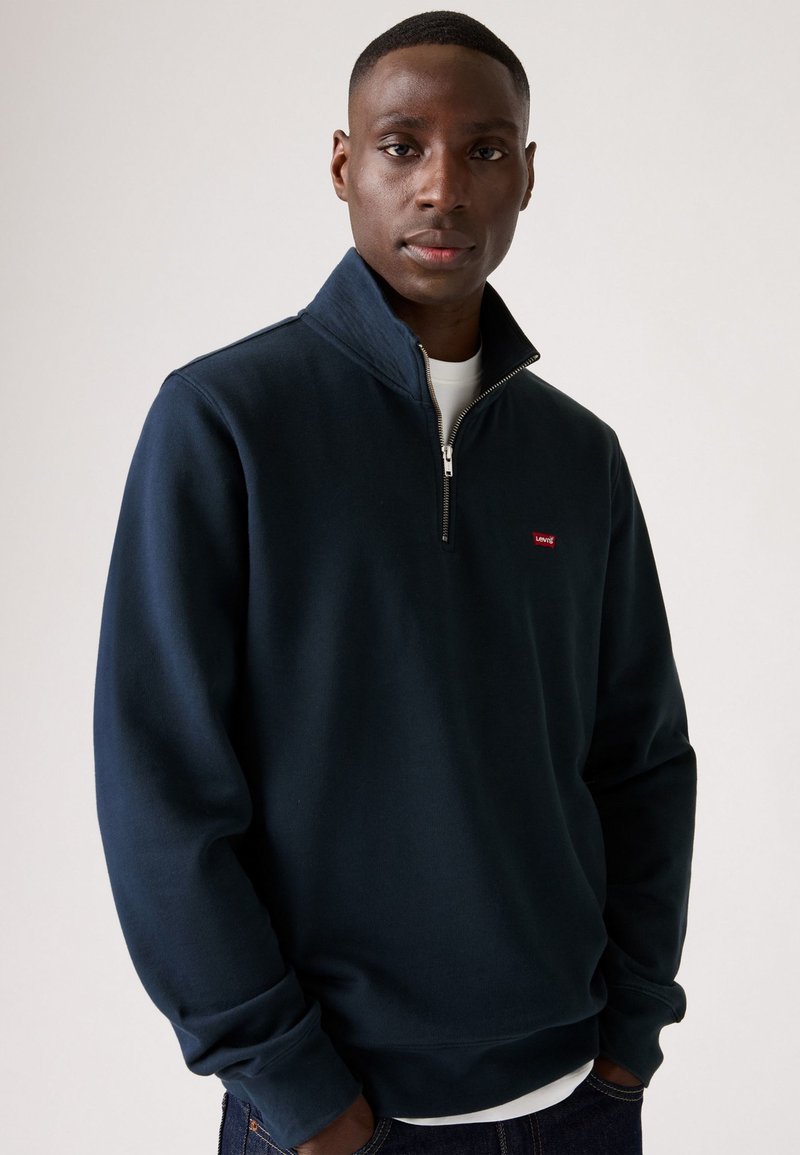 Donkerblauwe sweatshirt met een hoge kraag en een halve rits. Voorzien van elastische manchetbanden en een klein rood logo op de borst. Zacht, textuurstof.