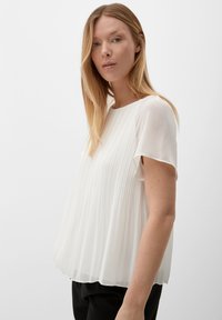 Blouse blanche plissée à manches courtes, fabriquée à partir d'un tissu léger. Elle présente un col rond et tombe légèrement des épaules.