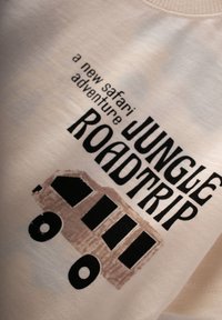 Heled kreem t-särk, millel on must ja pruun graafiline tekst ning bussijoonistus, millel on kirjas "JUNGLE ROADTRIP" ja "uus safariseiklus."