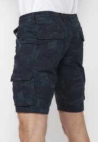 Marina cargo shorts med elastisk midja, flera fickor och ett strukturerat mönster. Tyget har en blandning av enfärgade och prickiga mönster.