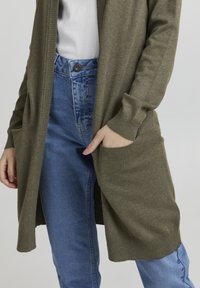 Olive green long cardigan with ribbed cuffs, worn over a white top and blue jeans. Features two front pockets for added detail.