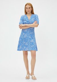 Korte mouwen blauwe jurk met wit bloempatroon, v-hals ontwerp, uitlopende rok en lichte stof. G gedragen met witte slippers.