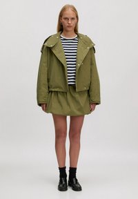 Olijfgroene oversized jas met grote kraag en knoopdetails; gestreepte marineblauwe en witte blouse; olijfgroene skaterskirt; zwarte instapper met sokken.