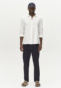 Bela srajca z dolgimi rokavi iz lanu, mornarsko modri hlače s stranskimi žepi, temna kapa in svetlo rjave sandale z nastavljivimi trakovi. Preprost, sproščen outfit.