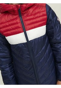 Vadderad jacka med en röd överdel, vit rand och marinblå kropp. Har dragkedja och huva, med en slät, glänsande yta.