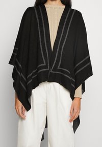 Svart poncho med grå ränder, tillverkad av mjukt tyg. Öppen frontdesign som hänger över axlarna. Bärs över en beige stickad topp och vita byxor.