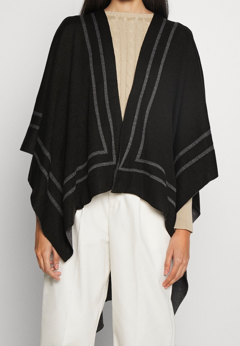 Svart poncho med grå ränder, tillverkad av mjukt tyg. Öppen frontdesign som hänger över axlarna. Bärs över en beige stickad topp och vita byxor.