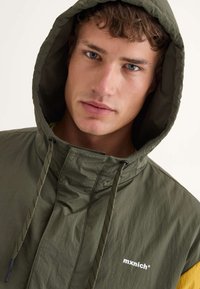 Chaqueta ligera verde oliva con capucha y cordones, y un pequeño logotipo blanco. Presenta un forro interior amarillo y una tela suave y texturizada.