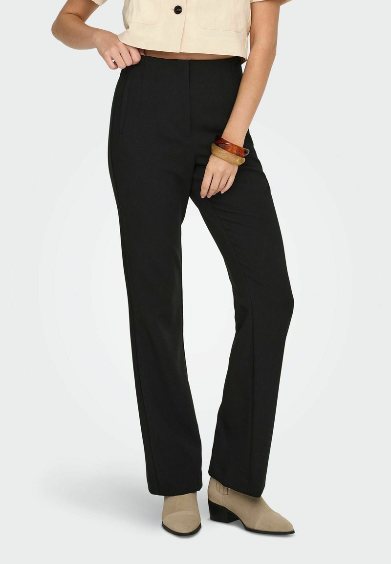 Pantalon noir taille haute à coupe ajustée, avec plis, associé à des bottines beiges. Texture de tissu lisse.