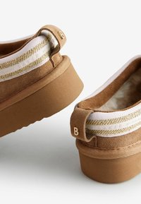 Brūni zamša slip-on apavi ar baltu joslu un zelta akcentiem, ar izturīgu zoli un cilpu uz papēža, kas marķēta ar "B". Mīksta iekšējā oderējuma.