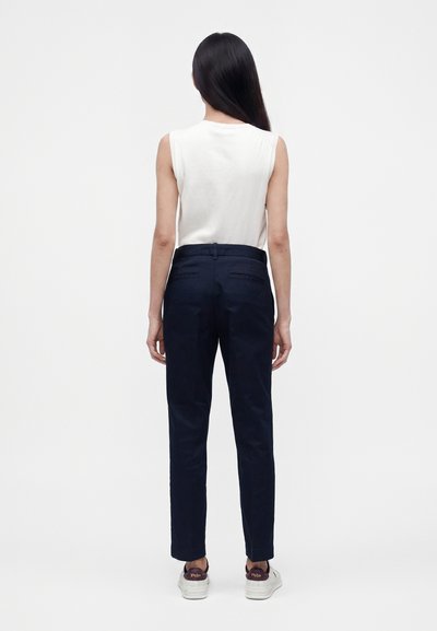 Femme aux longs cheveux noirs, debout de dos, portant un haut blanc sans manches, un pantalon bleu marine foncé et des baskets blanches avec des accents foncés.